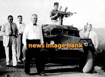 November 1957: Members of the American Tin Mission, escorted by an armoured vehicle, walk along the barbed wire fence at the Sungei Buloh Agricultural Resettlement Camp, Selangor. This is the second resettlement camp they visited during the day.